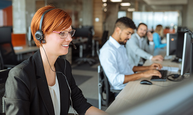 A smiling IT help desk employee wearing headphones sitting alongside her coworkers