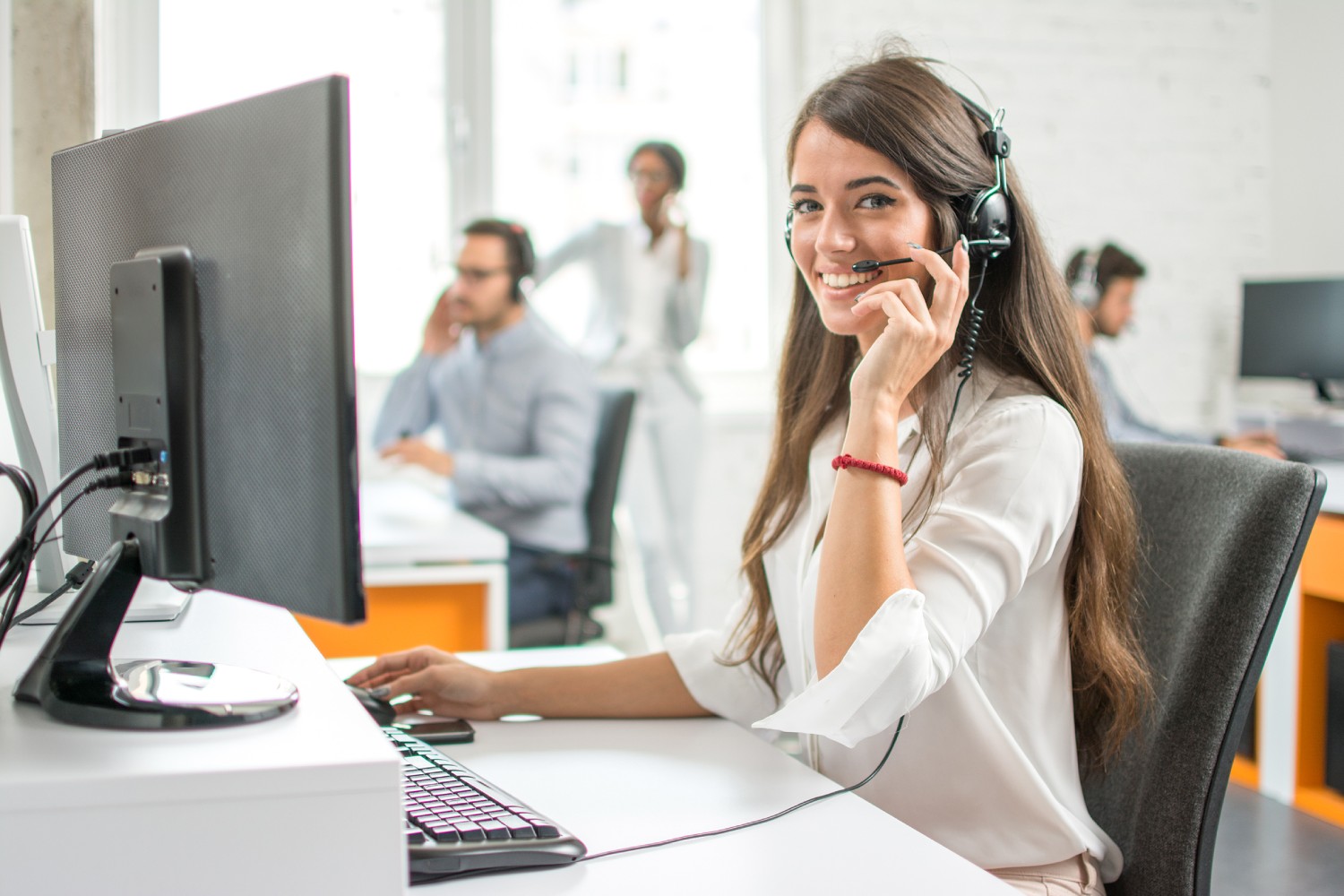 Young,Friendly,Operator,Woman,Agent,With,Headsets,Working,In,A A smiling IT help desk employee wearing headphones sitting alongside her coworkers